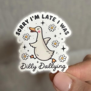 May include: A white sticker featuring a cartoon duck with an orange beak and feet, surrounded by daisies and stars. The text reads "Sorry I'm late I was Dilly Dallying." The sticker has a white border and is on a grey background.