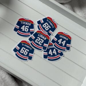 May include: A selection of hockey jersey stickers. Each sticker is a miniature replica of a hockey jersey, with names and numbers such as "Tucker 46", "Di Laurentis 66", "Logan 22", and "Graham 44". The jerseys are mainly navy blue with red and white detailing.