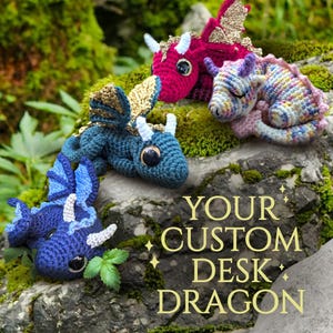 Op de afbeelding: Een verzameling handgemaakte gehaakte draken in verschillende kleuren, waaronder blauw, rood en paars, met gouden en witte accenten. De draken zijn geposeerd op een met mos bedekte rots, met de gouden tekst "YOUR CUSTOM DESK DRAGON".