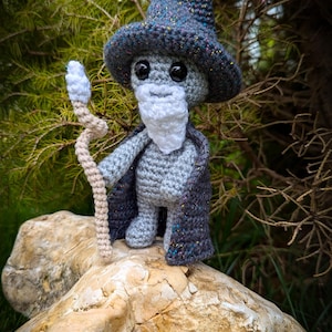 Peut inclure: Poupée de magicien au crochet faite à la main, avec un corps gris, un chapeau pointu et une longue barbe. Le magicien porte une cape bleu foncé et tient un bâton. La poupée est posée sur un gros rocher, avec un fond vert.