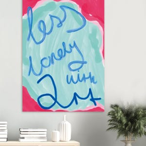 Peut inclure: Une œuvre d'art abstraite colorée avec les mots "less lonely with art" en bleu sur un fond turquoise, encadrée de rose et de blanc. L'œuvre est affichée sur un mur blanc.