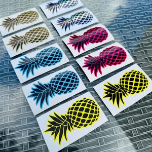 Op de afbeelding: Een verzameling ananasvormige stickers in verschillende kleuren, waaronder geel, blauw, roze en zilver. Elke sticker heeft een gedetailleerd rasterpatroon en een zwarte steel en bladeren. De stickers zijn gerangschikt op een getextureerd oppervlak.