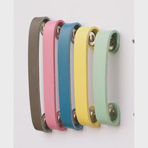 May include: A set of five leather cabinet pulls in a row, each a different color: gray, pink, blue, yellow, and green. The pulls are attached to a white surface with silver hardware. The leather straps are curved.