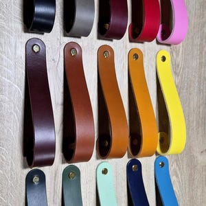 May include: A selection of leather drawer pulls in a range of colours: black, grey, red, pink, brown, orange, yellow, green, blue, and teal. Each pull is a loop with a brass rivet.