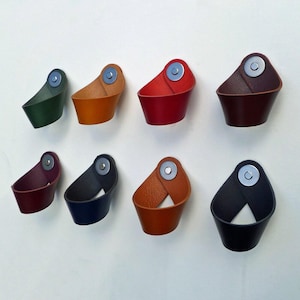 May include: A collection of eight leather wall hooks in various colors, including green, orange, red, and black. Each hook features a curved design with a metal disc accent, ideal for hanging items.