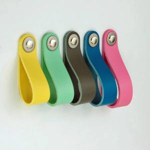 May include: Five colourful leather drawer pulls in yellow, mint green, brown, blue, and pink. Each pull is attached with a silver rivet. The pulls are arranged in a row against a white background.