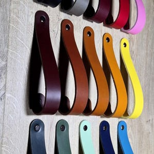 May include: A collection of leather drawer pulls in various colors, including black, gray, burgundy, red, pink, brown, orange, yellow, green, mint, navy, and blue. Each pull is a loop shape with a rivet at the top, mounted on a wooden surface.
