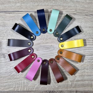 May include: A selection of leather pull tabs in various colours, each with a brass rivet. The tabs are arranged in a circle, displaying shades such as black, blue, green, yellow, and brown. These leather accessories are suitable for drawers or cabinets.
