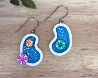 May include: A pair of novelty earrings in the shape of swimming pools. Each earring has a blue pool design with a white border, and includes miniature pool toys such as a beach ball, a life preserver, and a pink and white striped lollipop.