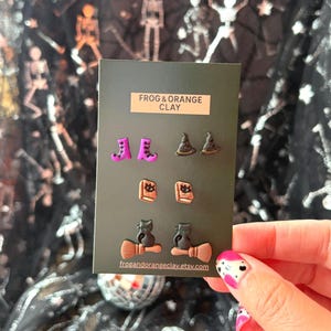 May include: A set of three pairs of stud earrings on a black display card labelled FROG & ORANGE CLAY. The earrings feature purple boots, black glittery witch hats, tan spellbooks with black symbols, and black cats sitting on tan broomsticks.