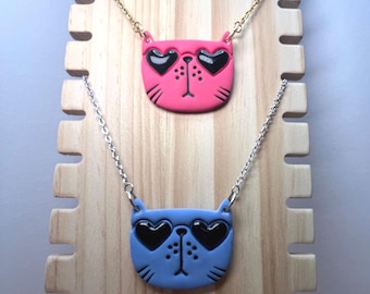 May include: Two necklaces featuring cat-shaped pendants. One pendant is pink, the other is blue. Both have black heart-shaped sunglasses and a small nose. The necklaces are displayed on a wooden stand.