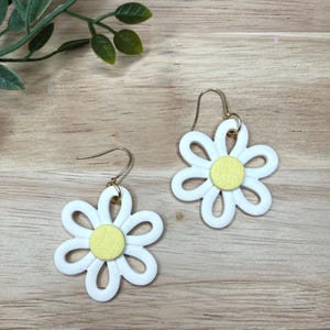 Könnte beinhalten: Ein Paar ohrringförmige Ohrringe. Jeder Ohrring hat eine weiße Blume mit gelber Mitte und goldfarbenen Ohrhaken. Die Ohrringe sind auf einer hellbraunen Holzoberfläche platziert.