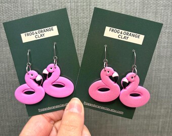 May include: A pair of pink flamingo earrings with black beaks and silver hooks. The earrings are presented on dark green cards, with the text "FROG & ORANGE CLAY" and a website address.