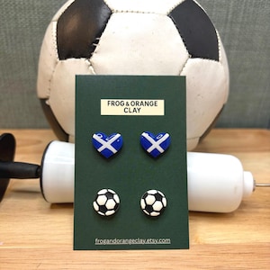 May include: A dark green card shows four clay earrings. Two are heart-shaped with a blue and white Scottish flag design. The other two are miniature footballs. The text "FROG & ORANGE CLAY" is at the top of the card.