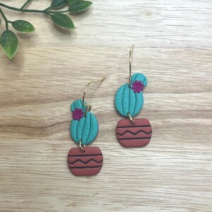 Pode incluir: Brincos feitos à mão com um design de cacto turquesa, uma pequena flor rosa e um vaso cor de terracota com listras pretas. Os brincos penduram-se em argolas douradas.