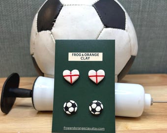 May include: A dark green card shows four clay earrings: two white hearts with red crosses and two miniature footballs. The card has the text "FROG & ORANGE CLAY" and the website address frogandorangeclay.etsy.com.