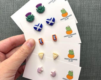 May include: A selection of handmade clay earrings on white cards. The earrings feature various designs, including thistles, heart-shaped flags, and miniature soda cans with "IRN" and "BRU" text. The cards are labelled "FROG & ORANGE CLAY".
