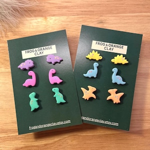 Può includere: Due biglietti verde scuro con figure di argilla colorate a forma di dinosauro. I biglietti presentano il testo "FROG & ORANGE CLAY" e l'indirizzo del sito web "frogandorangeclay.etsy.com". I dinosauri sono nei toni del viola, rosa, verde, giallo e arancione.