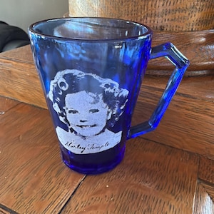 May include: A cobalt blue glass mug featuring a portrait of Shirley Temple. The mug has a unique angular handle and the name "Shirley Temple" is printed below the portrait. The mug is sitting on a wooden surface.