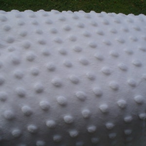 May include: A close-up of a white, textured blanket. The blanket has a soft, plush appearance with a pattern of evenly spaced, raised, circular dots. The background is a blurred view of green grass.
