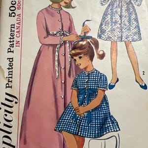 May include: Vintage Simplicity printed sewing pattern for a child's dress and robe. The illustration features three girls in dresses and robes. The pattern includes a pink robe, a blue and white gingham dress, and a floral patterned robe.