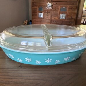 May include: An oval turquoise glass casserole dish with a white snowflake pattern around the exterior. The dish includes a clear glass lid with a central handle and integrated side handles on the base for easy lifting and transport.