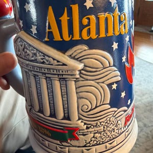 May include: A white ceramic mug with a black handle and the text "But First, Coffee" in a bold, black font. The mug is set against a blurred background, suggesting a cozy setting.