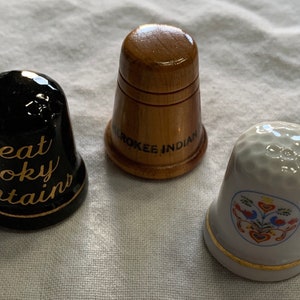 May include: Three thimbles, one black with gold lettering that reads "Great Smoky Mountains", one wooden with black lettering that reads "Cherokee Indian", and one white with a colorful floral design.