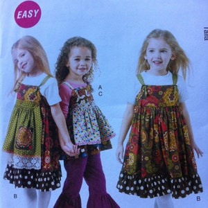 May include: Three girls wearing different patterned dresses with ruffled hems and polka dot trim. The dresses are all sleeveless and have a white top with straps. The girls are wearing white tights and shoes. The image is from a McCall's pattern for easy sewing.