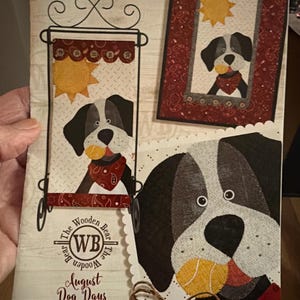 May include: A brochure for "Little Quilts Monthly Table Toppers" featuring a dog-themed design. The cover includes a dog graphic with a red bandana and a yellow ball, along with the text "August Dog Days" and "The Wooden Bear".