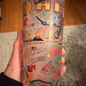 May include: A frosted glass featuring a design with the word "Ohio" in gold, along with illustrations and text including "Lake Erie", "Toledo", "Columbus", and "Reds". The glass is cylindrical and appears to be held by a person.