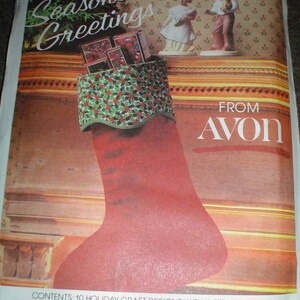Pu&ograve; includere: Un calzino rosso con un polsino a motivo di agrifoglio verde e bianco pieno di piccole scatole. Il calzino &egrave; appeso a un camino con un bordo decorativo. Il testo "Season's Greetings" &egrave; scritto in un carattere festivo sopra il calzino. Il testo "FROM AVON" &egrave; scritto in un carattere grande sotto il calzino. Il testo "CONTENTS: 10 HOLIDAY CRAFT DESIGNS WITH INSTRUCTIONS FSC 5380 AVON PRODUCTS INC., DISTR. NEW YORK, NEW YORK 10019" &egrave; scritto in fondo all'immagine.