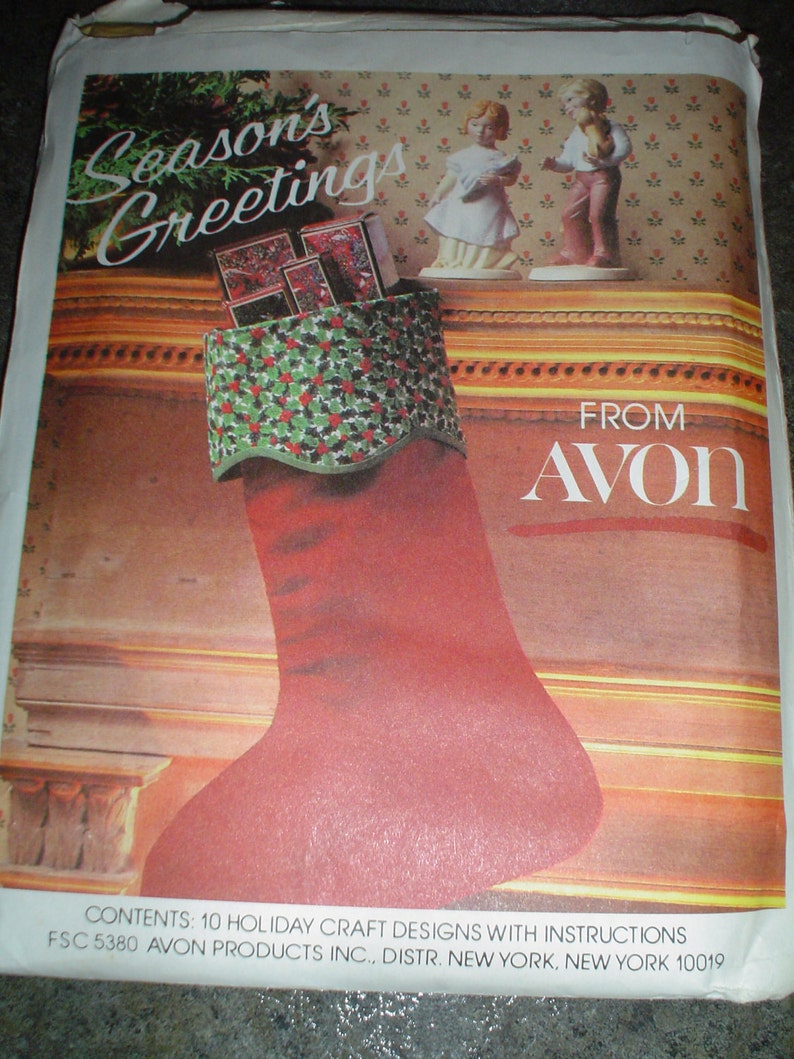 Pu&ograve; includere: Un calzino rosso con un polsino a motivo di agrifoglio verde e bianco pieno di piccole scatole. Il calzino &egrave; appeso a un camino con un bordo decorativo. Il testo "Season's Greetings" &egrave; scritto in un carattere festivo sopra il calzino. Il testo "FROM AVON" &egrave; scritto in un carattere grande sotto il calzino. Il testo "CONTENTS: 10 HOLIDAY CRAFT DESIGNS WITH INSTRUCTIONS FSC 5380 AVON PRODUCTS INC., DISTR. NEW YORK, NEW YORK 10019" &egrave; scritto in fondo all'immagine.