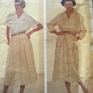 May include: A Vogue Patterns American Designer Calvin Klein sewing pattern envelope, number 1320. The front displays two illustrations of a light-colored, collared shirt and a pleated midi skirt, both cinched at the waist with a thin brown belt.
