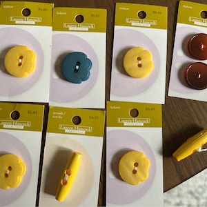 May include: Assortment of yellow, blue, and brown plastic buttons and toggles on white cards with the text "Lauren Hancock Collection" and prices.