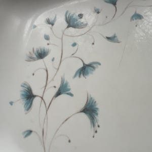 May include: A white ceramic plate with a blue and brown floral design. The flowers are stylized and have a delicate, whimsical look.