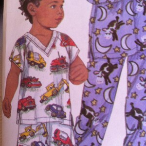 May include: A child's pajama set featuring a white top and shorts printed with colorful construction vehicles. The matching pajama pants are purple with a pattern of cows, moons, and stars. The top has a V-neck.