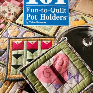 May include: A book titled "101 Fun-to-Quilt Pot Holders" by Trice Boerens, with a blue cover. The image showcases various quilted pot holders in different colors and patterns, including floral and geometric designs. The pot holders are square and rectangular.