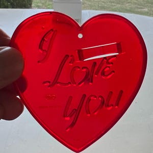 May include: Red plastic heart-shaped ornament with the words "I Love You" inscribed on it. The ornament has a small hole at the top for hanging.