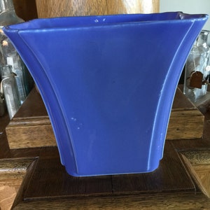 May include: A blue ceramic vase with a square shape and a wide opening. The vase has a smooth, glossy finish and is sitting on a wooden surface.