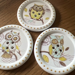 May include: Three round wooden buttons with a white background and a gold polka dot border. Each button features a different owl design with brown, yellow, and blue accents.