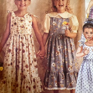 May include: Two young girls in vintage floral dresses. One dress is cream-colored with brown and yellow flowers, the other is blue with white and yellow flowers. The girls are holding hands and smiling. The dresses have ruffles and bows.
