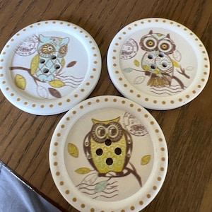 May include: Three white wooden buttons with brown polka dots around the edge. Each button features a different owl design with brown, yellow, and blue accents. The owls are perched on branches with leaves.