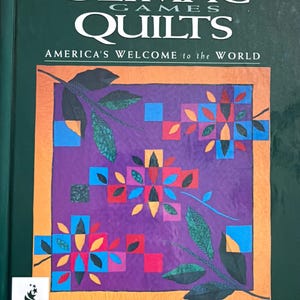 May include: A book cover titled "The Olympic Games Quilts" with the subtitle "America's Welcome to the World." The cover features a purple quilt design with orange, blue, red, and yellow geometric shapes, and green leaf accents. The Atlanta 1996 logo is visible.
