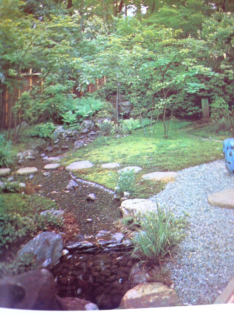 May include: A Japanese garden with a small stream running through it. The stream is lined with rocks and there are stepping stones leading across it. The garden is covered in green moss and there are trees and bushes in the background.
