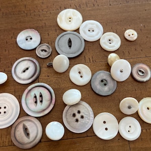 May include: A selection of vintage buttons in white, cream, and grey hues. The buttons are of different sizes and designs, including round, oval, and flat styles with two or four holes. Some buttons have a pearlescent finish.