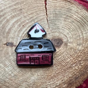 May include: A ceramic button shaped like a red house with a white roof and black trim. The house has two windows and a door. The button has two holes for sewing.