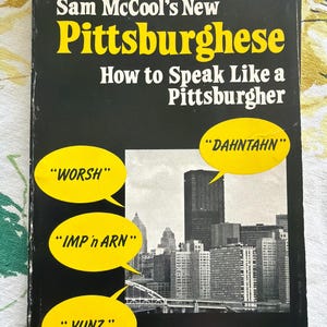 May include: A book titled "Sam McCool's New Pittsburghese: How to Speak Like a Pittsburgher." The cover is black with yellow and white text. Speech bubbles with phrases like "WORSH" and "DAHNTAHN" are featured. A black and white cityscape is in the background.