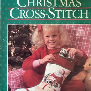 May include: A vintage "Better Homes and Gardens" Christmas Cross-Stitch book cover. The cover features a young girl holding a gift, with a Christmas stocking in the foreground. The stocking is decorated with a Santa Claus design and other festive elements.