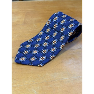 May include: A blue necktie featuring a repeating geometric pattern of black diamonds containing smaller, multi-colored star-like shapes in shades of yellow, white, and red. The fabric has a visible diagonal weave texture throughout the entire surface.